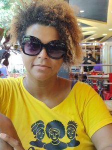 Nina Fola é militante do movimento negro, socióloga, produtora cultural e doutoranda em sociologia. Atualmente é a liderança negra que coordena o Coletivo Atinúké. Ela é uma mulher negra de cabelo crespo com luzes loiras e na foto está de óculos de sol e camiseta amarela. 